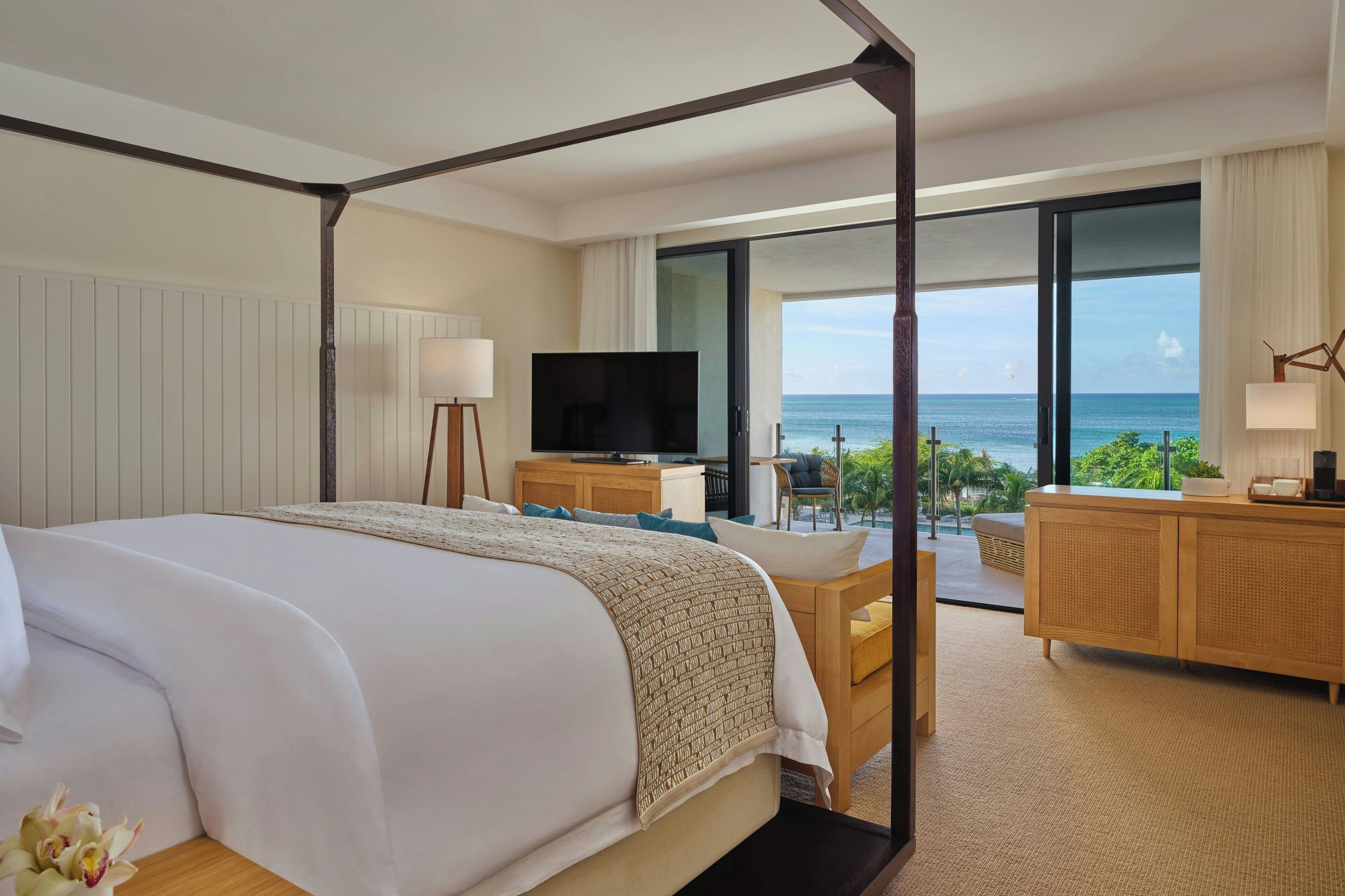 Quarto de hotel com cama de dossel, televisão, escrivaninha com cadeira, armário de madeira e vista para o oceano.