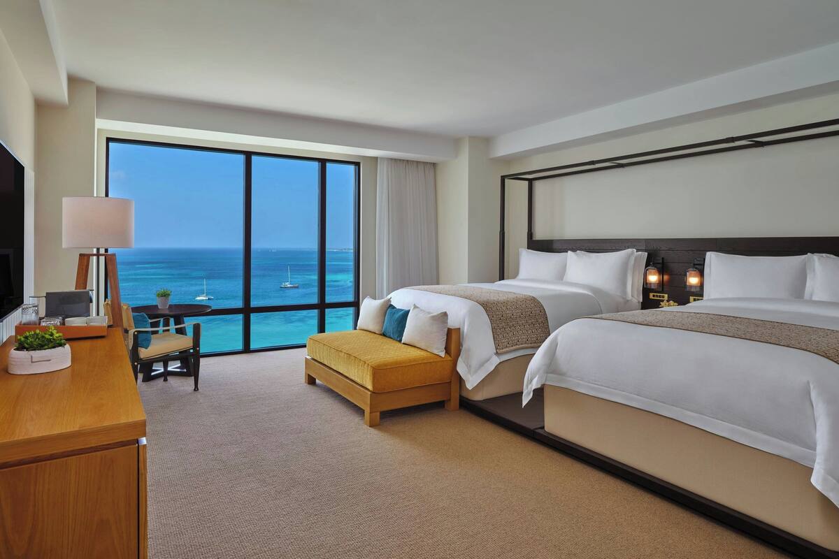 Hotelkamer met groot raam en uitzicht op zee, een bed, bureau met lamp en een klein zithoekje.