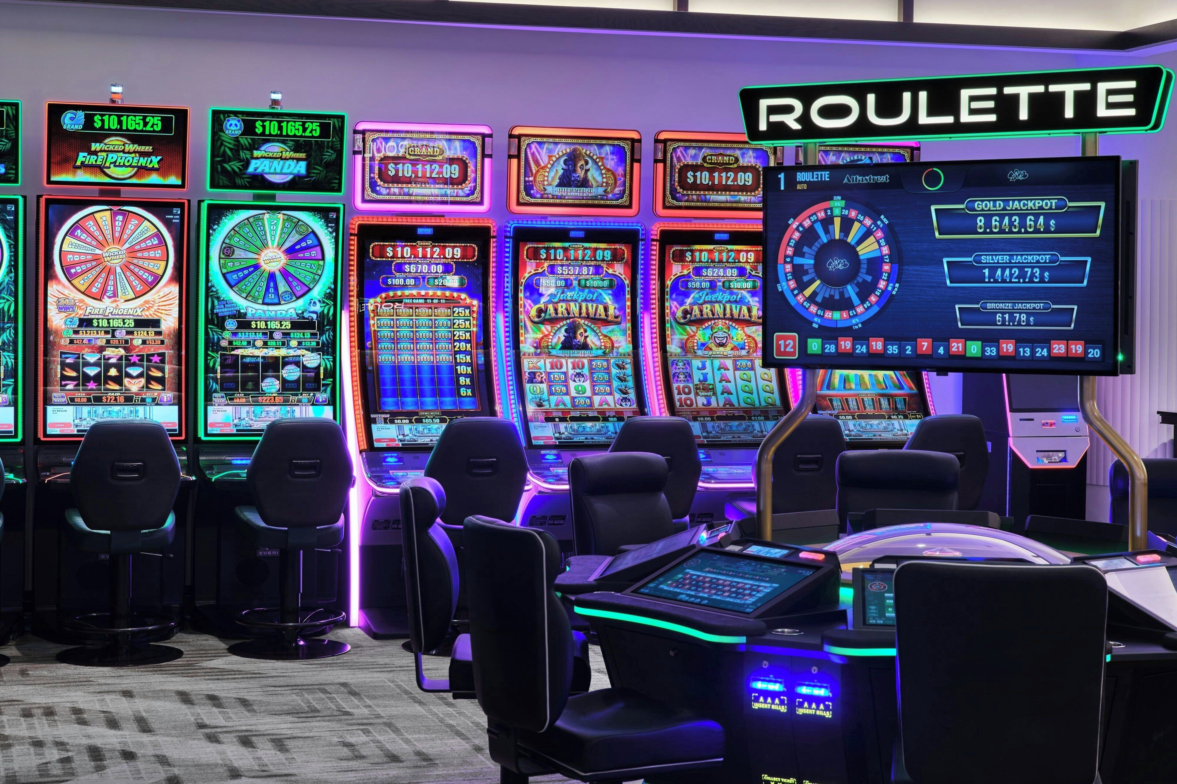 Un espace de jeux comprenant de nombreux appareils à sous et une table de roulette, avec différents thèmes de jeux et un grand écran affichant les montants des jackpots.