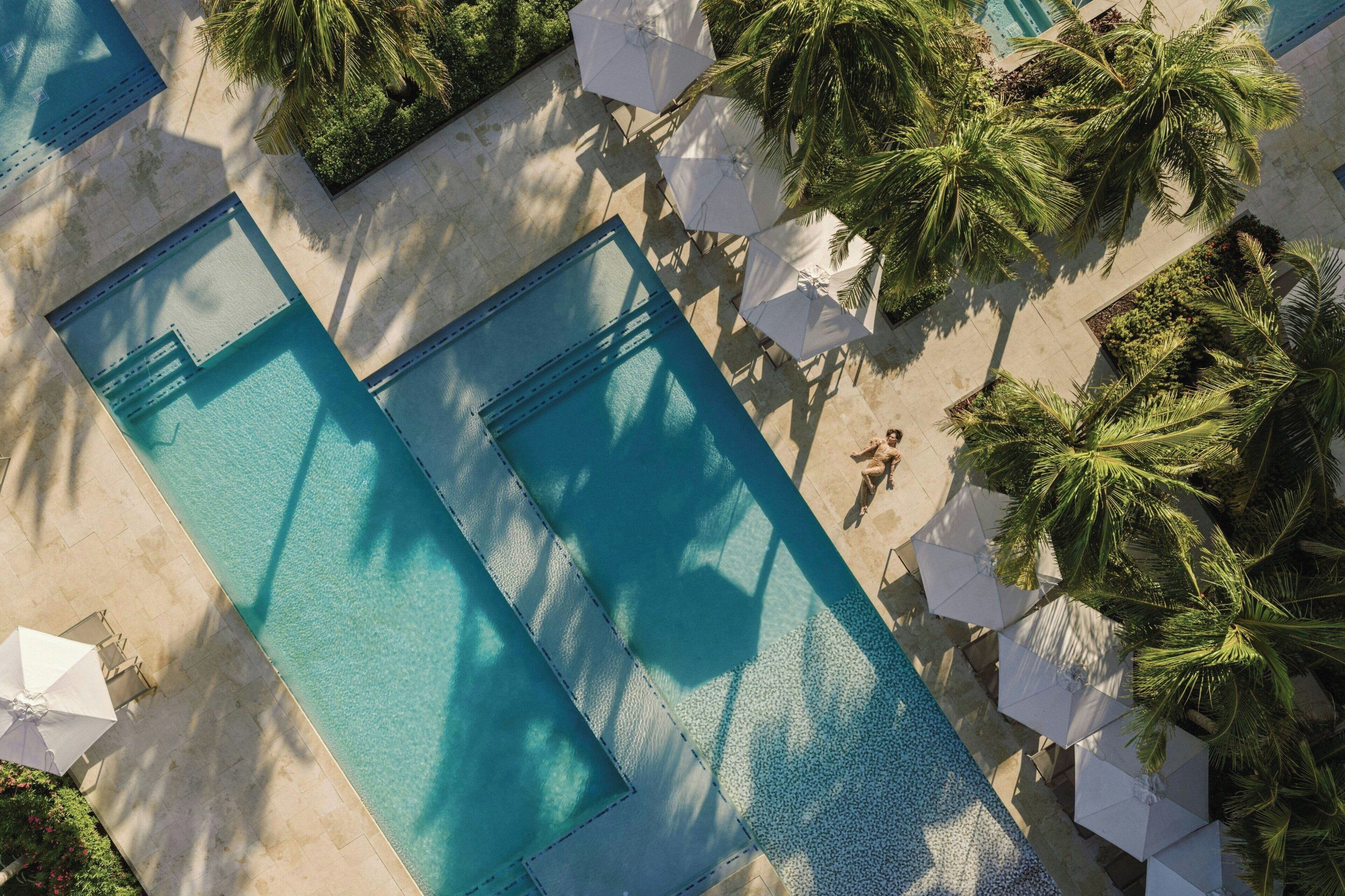 Vista aérea de uma área de piscina com várias piscinas, guarda-sóis brancos e palmeiras.