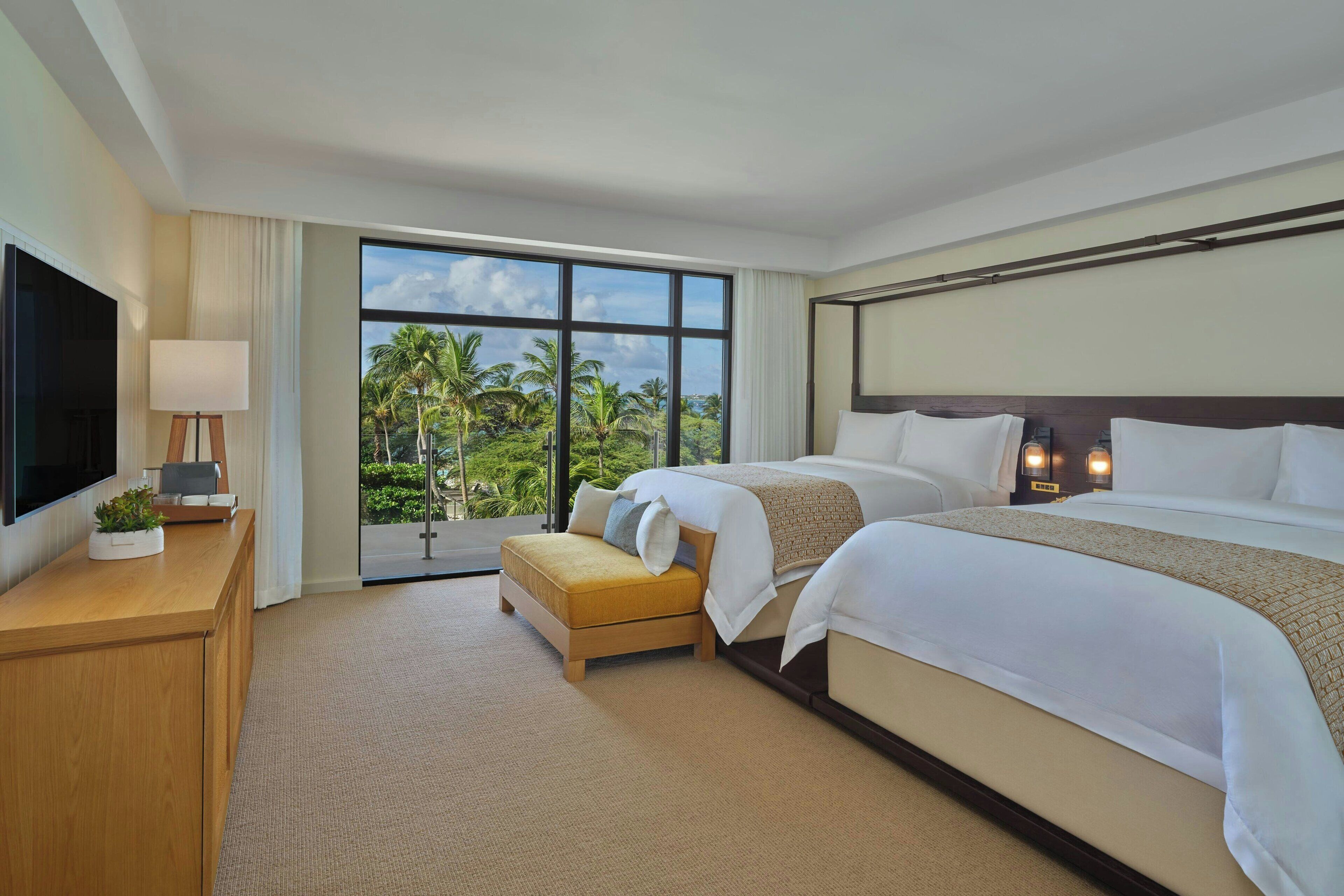 Habitación de hotel con dos camas, televisión, escritorio con lámpara y vistas a palmeras.