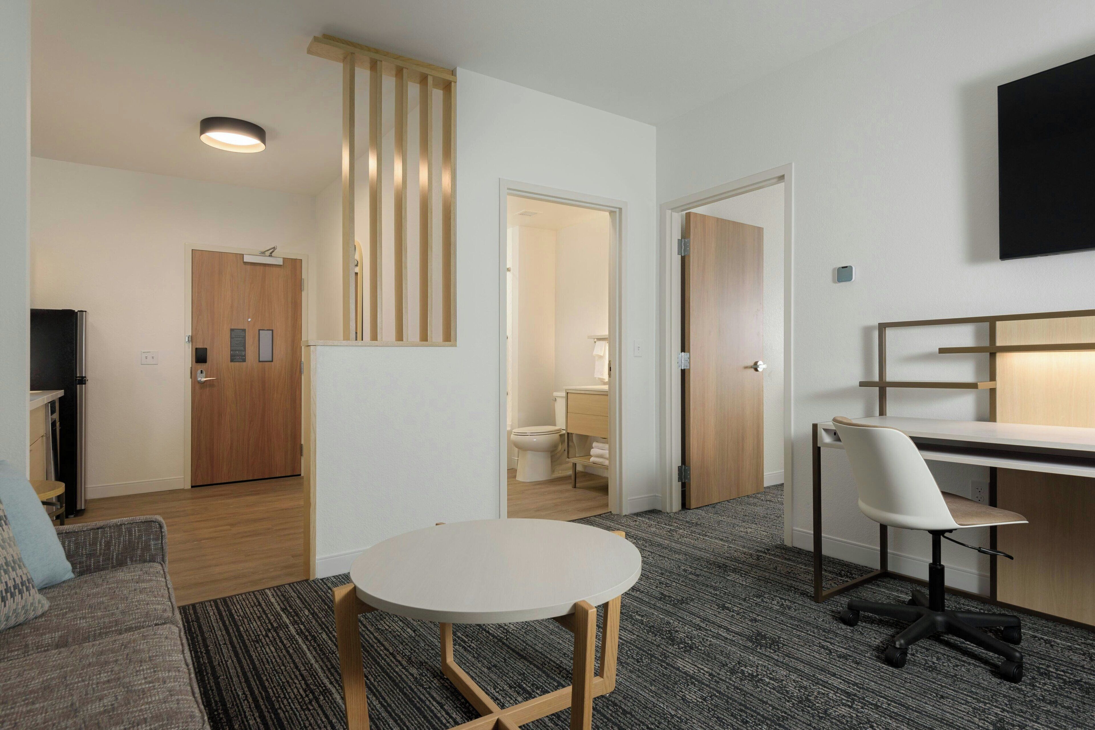 Une chambre d’hôtel moderne avec une table ronde, un canapé, un bureau avec une chaise, un téléviseur fixé au mur et une porte en bois.