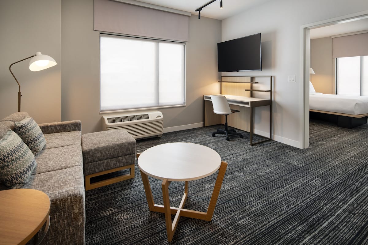 Een moderne hotelkamer met een bank, een ronde tafel, een bureau met stoel, een flatscreen-tv en een groot raam met jaloezieën.