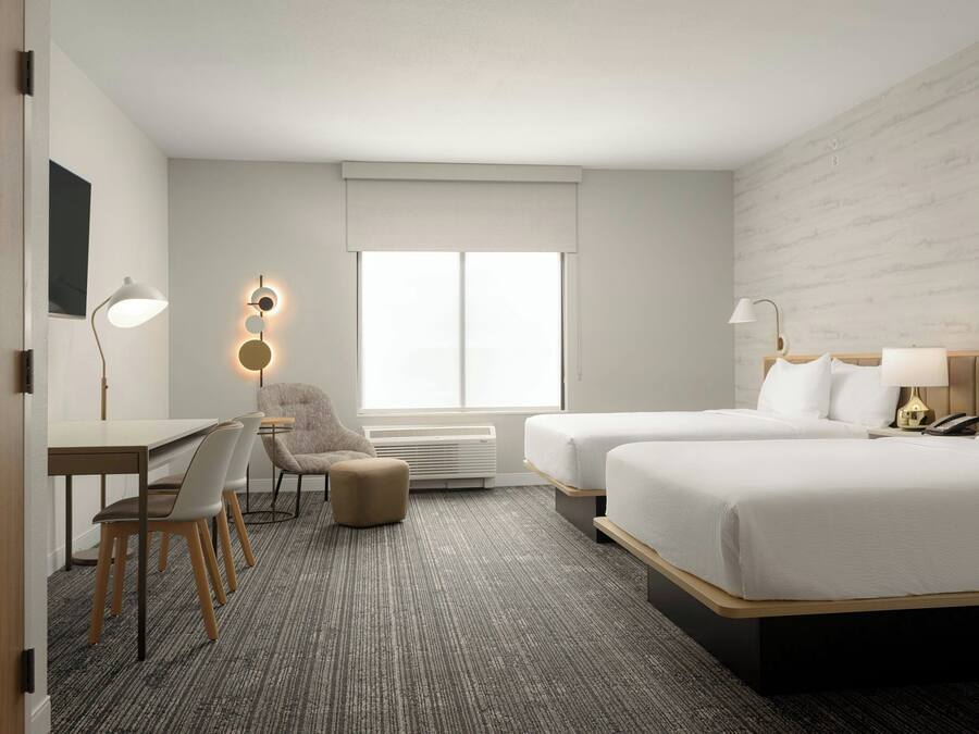 A hotel room with two beds, a desk, a chair, a lamp, and a large window.
