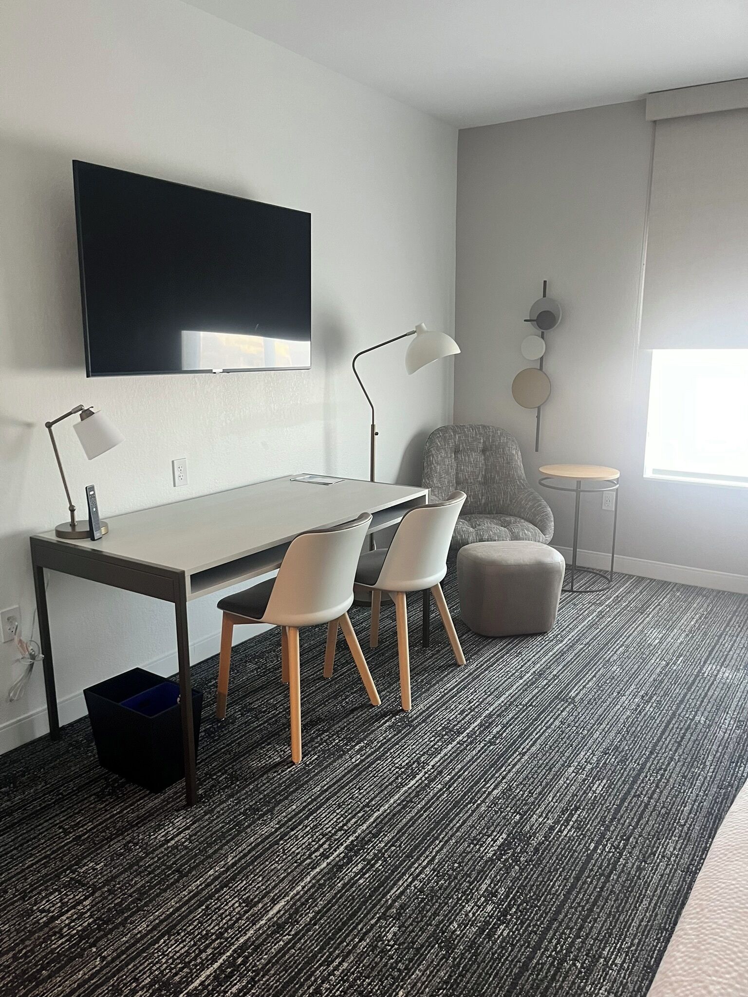 A modern hotel room with a flat-screen TV mounted on the wall, a desk with two chairs, a small round table, and a grey armchair.