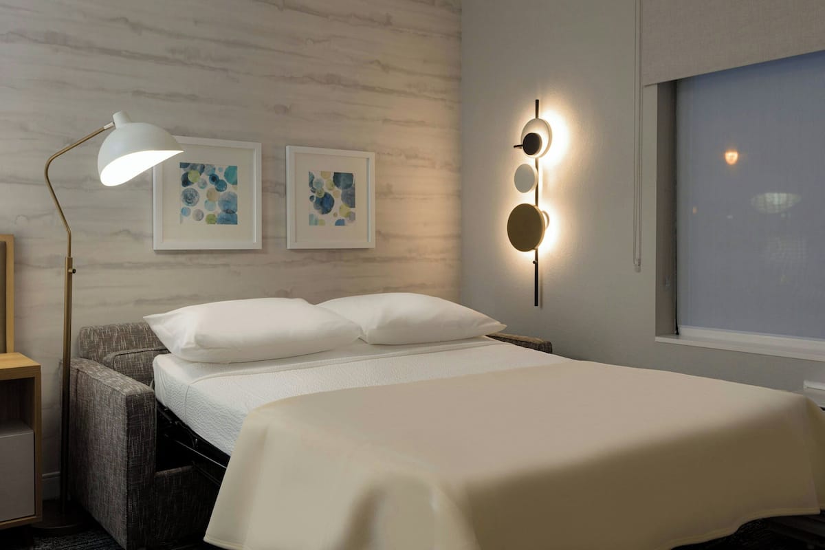Een keurig opgemaakt bed met witte lakens, een bedlamp en twee ingelijste abstracte kunstwerken aan de muur.