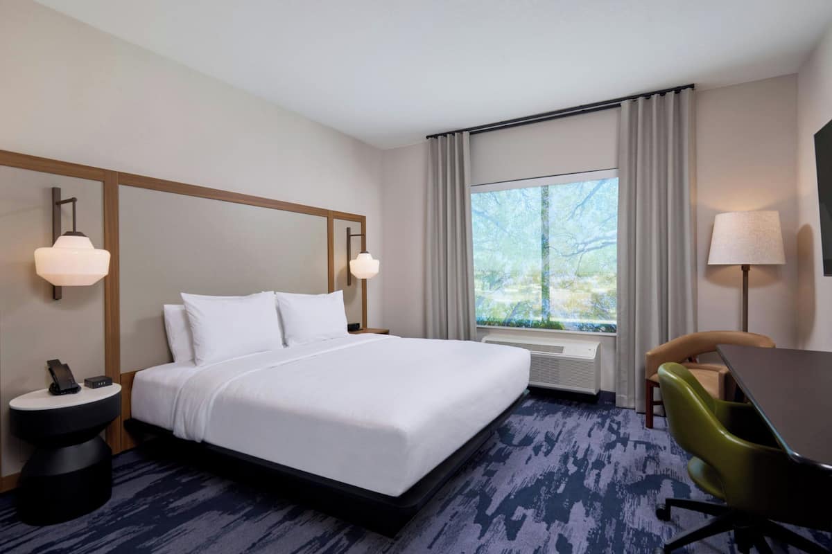 Une chambre d’hôtel avec un grand lit, un bureau, une chaise, une lampe, un téléphone sur une table de chevet et une fenêtre avec des rideaux.