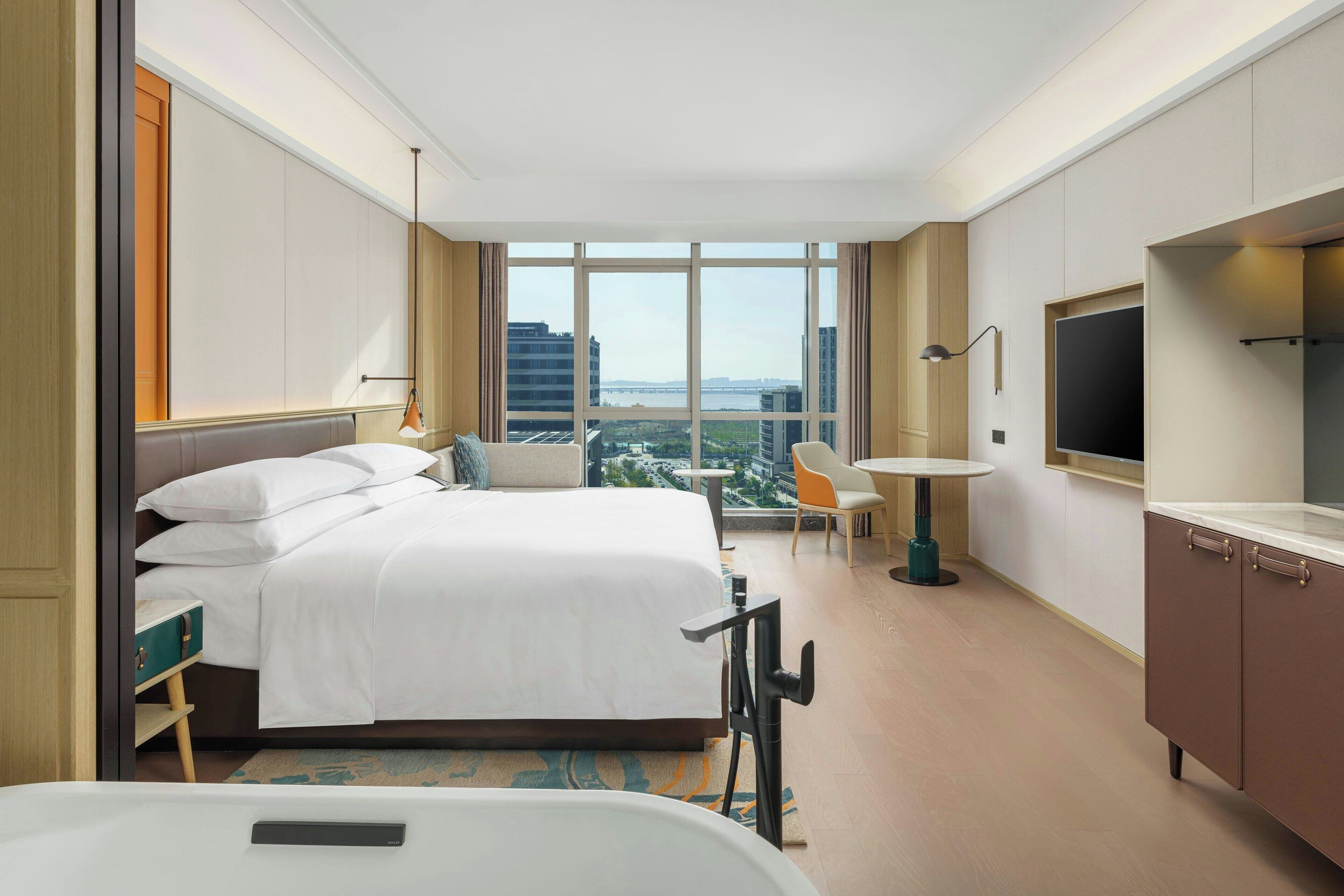 A hotel room with a large bed, a desk, a chair, a TV, and a view of the city through large windows.