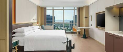 Ein Hotelzimmer mit einem großen Bett, einem Schreibtisch, einem Sessel, einem Fernseher und Blick auf die Stadt durch große Fenster.