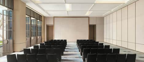 Un salón de conferencias con filas de sillas negras, una alfombra estampada y un ventanal grande en un lateral.