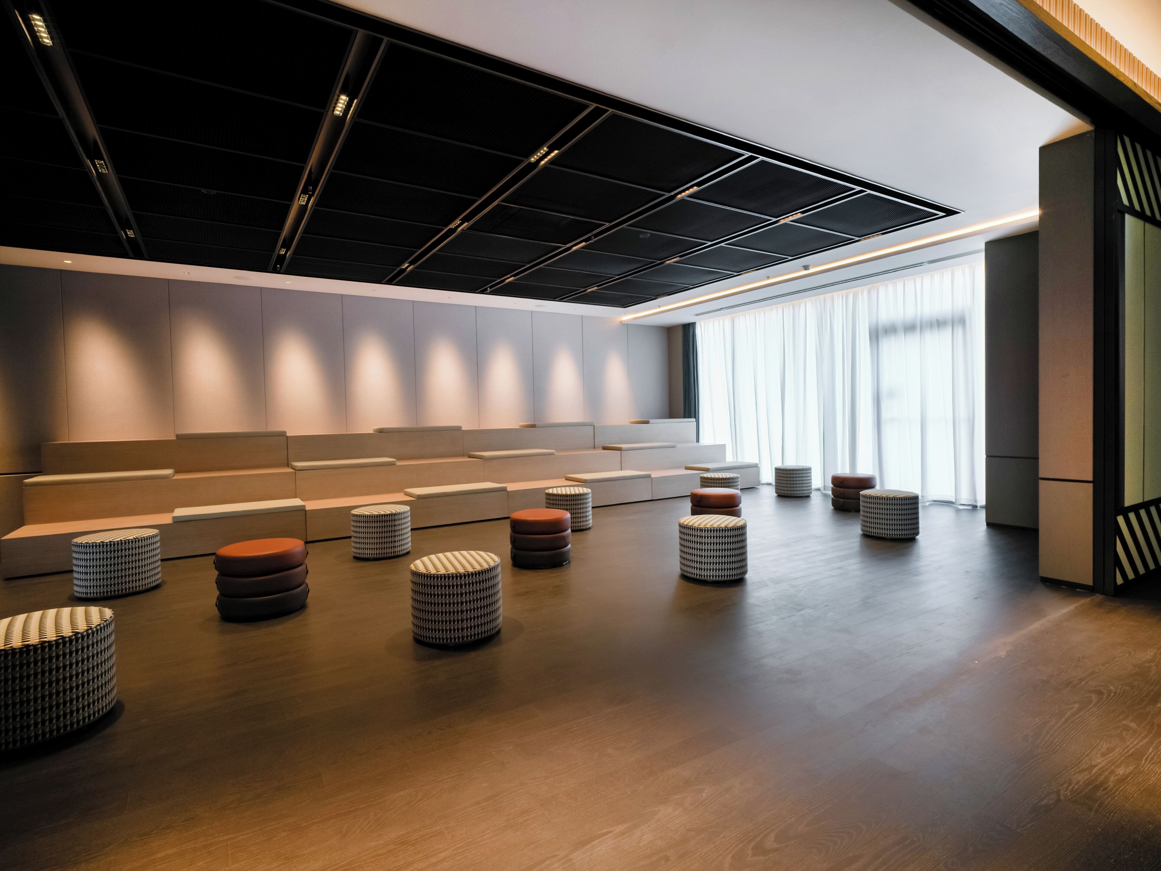 A modern interior with tiered seating and round ottomans, a wooden floor, and a large window with sheer curtains.