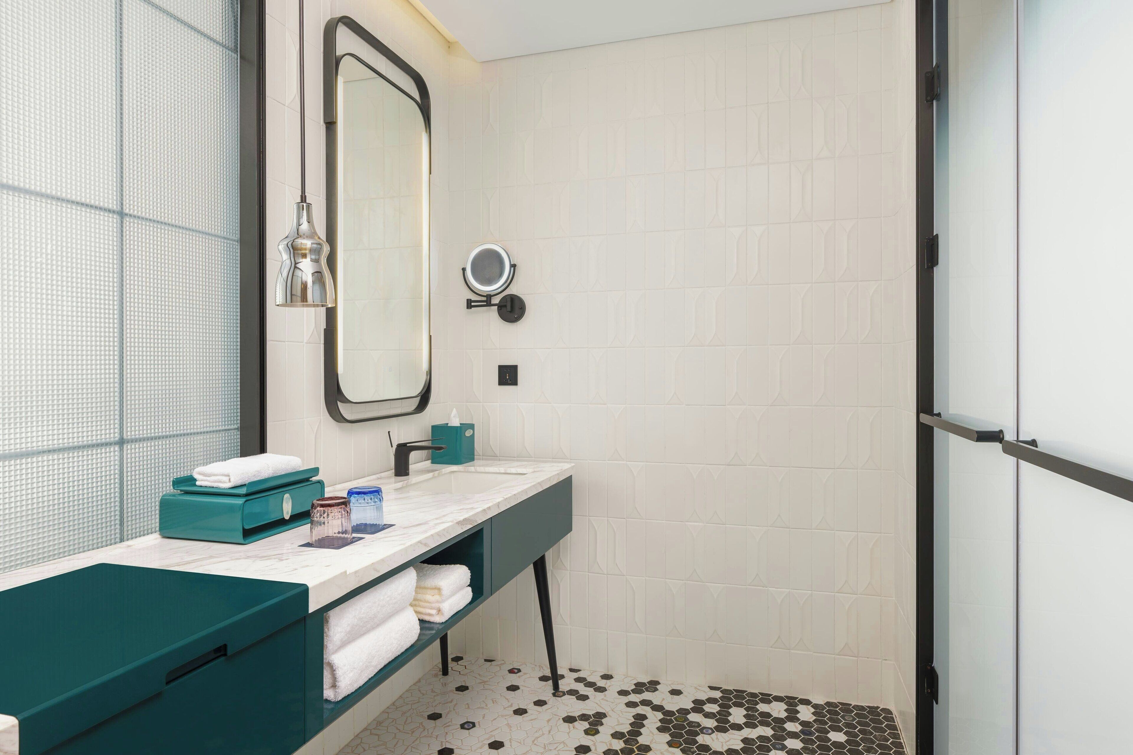 A modern bathroom with a white tiled wall, a large mirror, a sink with a teal countertop, and a shower area.