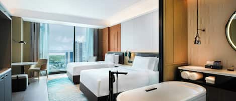 A modern hotel room with two beds, a sitting area, a large window with a city view, a bathtub, and a bathroom with a mirror and towel rack.