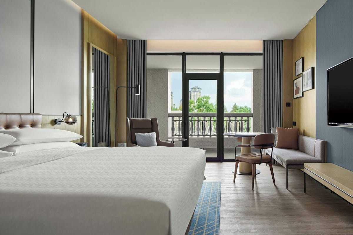 Une chambre d’hôtel avec un grand lit, un coin salon avec des fauteuils et une table, une télévision et un balcon avec vue.