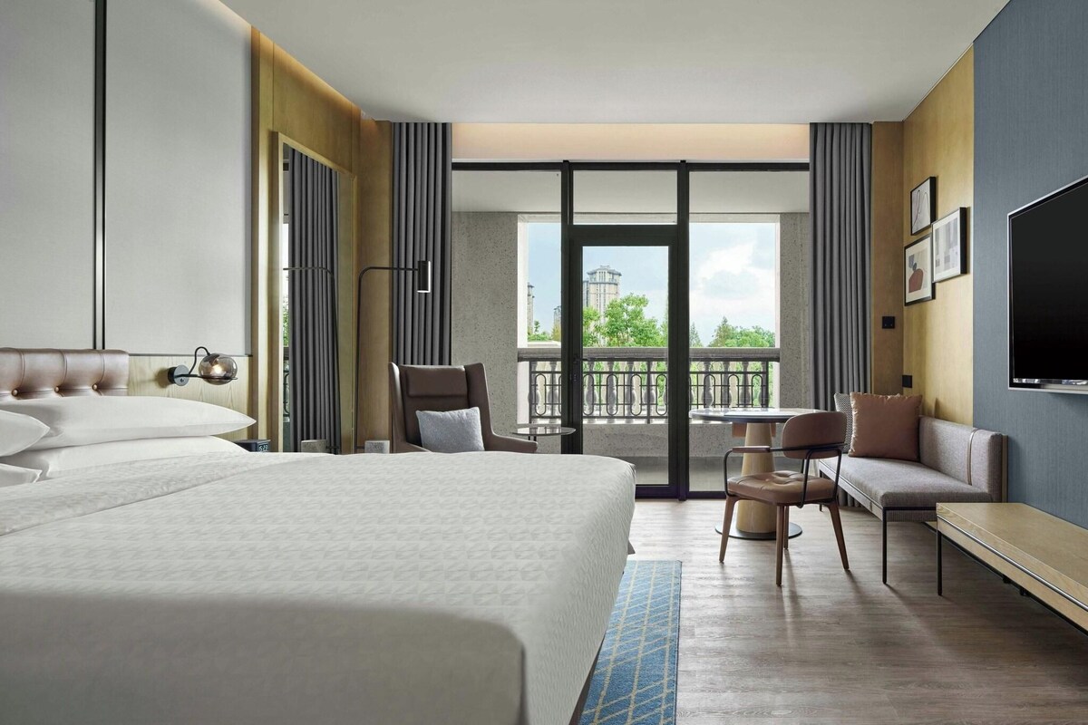 A hotel room with a large bed, a seating area with chairs and a table, a TV, and a balcony with a view.