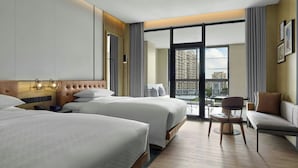 A hotel room with two beds, a sofa, a chair, a small table, and a balcony with a view of buildings.