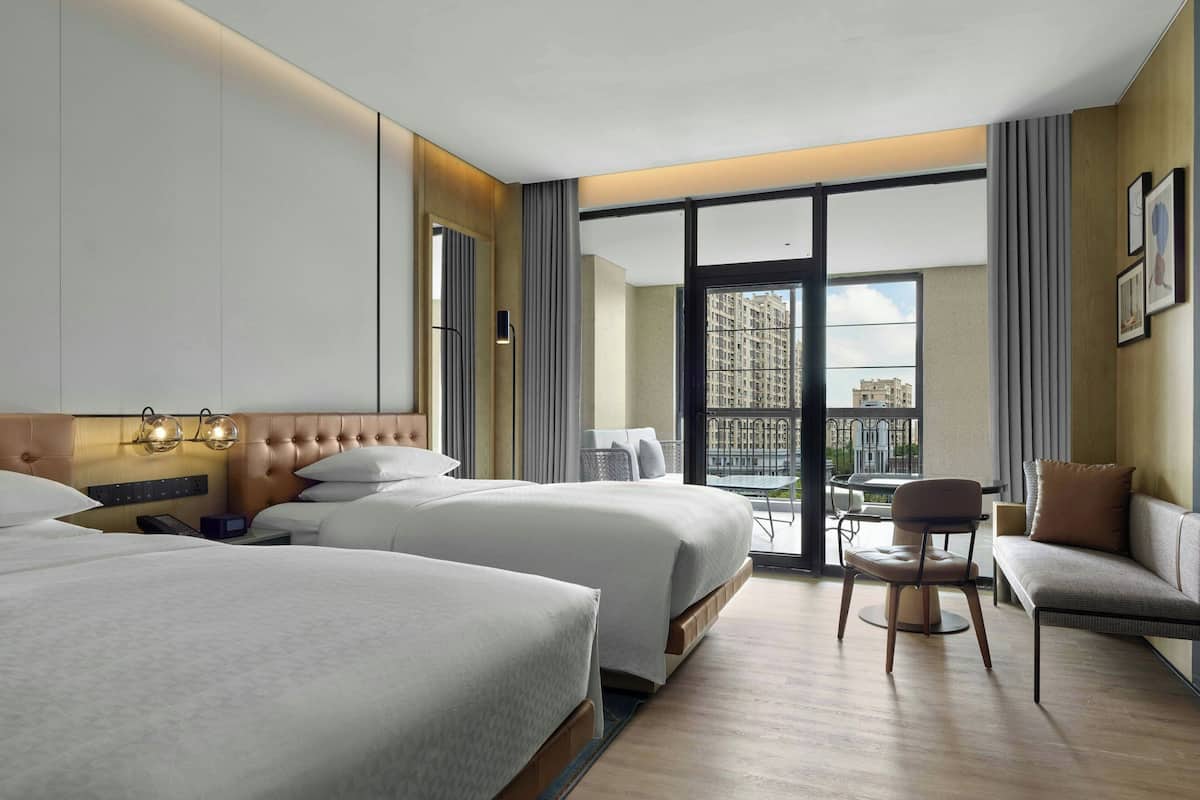 Une chambre d’hôtel avec deux lits, un canapé, une chaise, une petite table et un balcon donnant sur des immeubles.