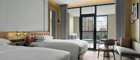 A hotel room with two beds, a sofa, a chair, a small table, and a balcony with a view of buildings.