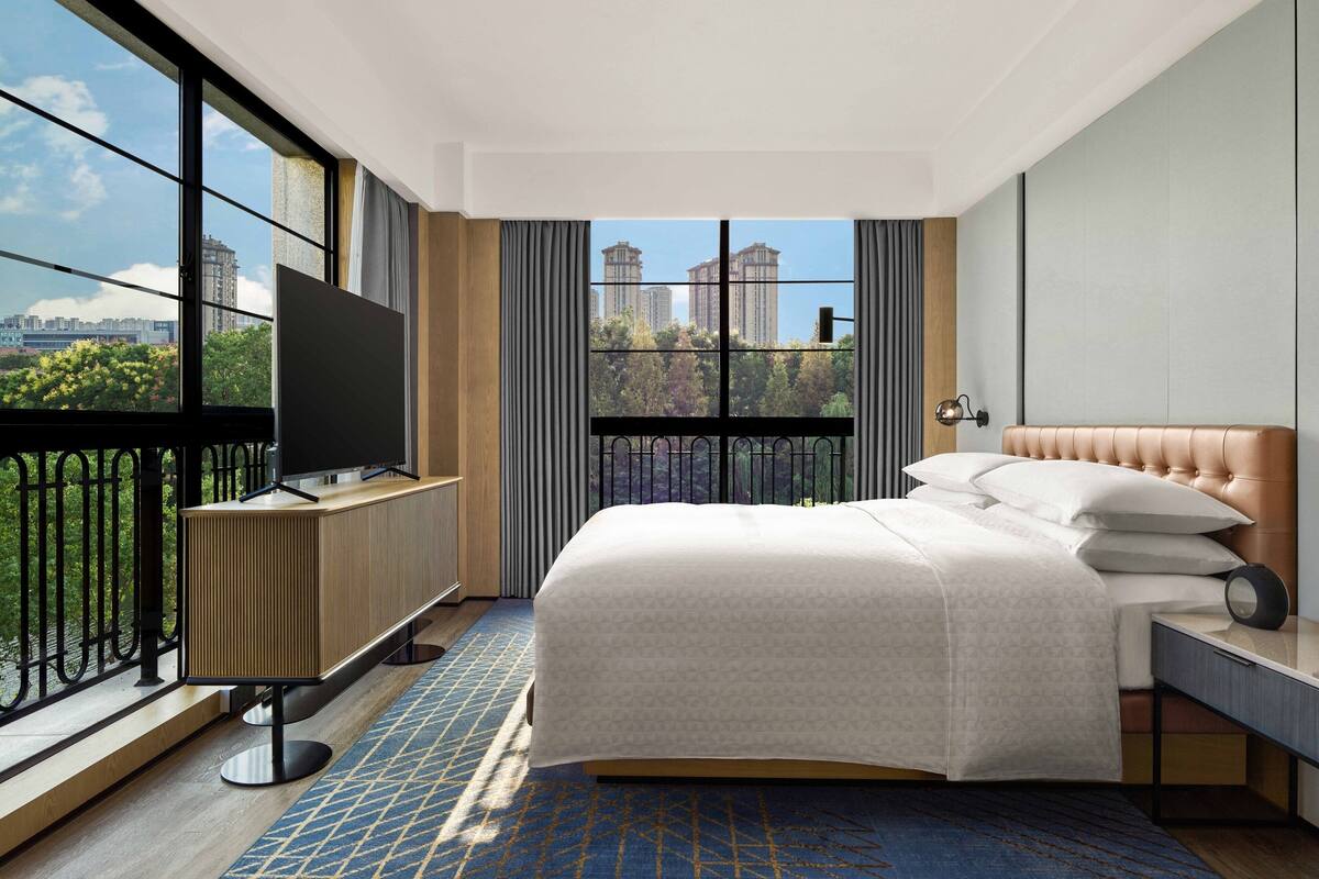 Une chambre d’hôtel moderne avec un grand lit, un téléviseur posé sur un meuble et un balcon offrant une vue sur la ville.
