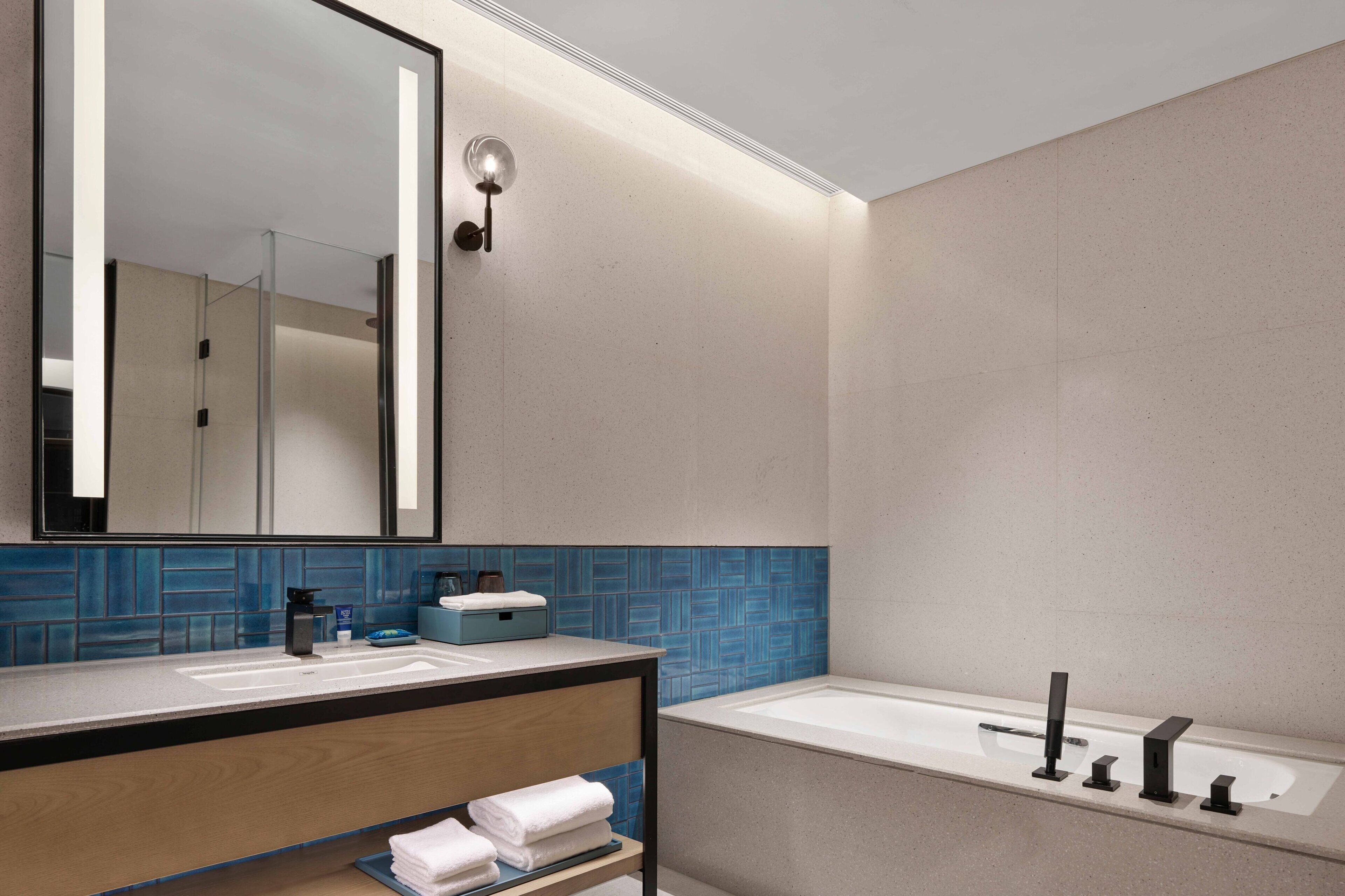 A modern bathroom with a large mirror, a sink, a bathtub, and a shelf with towels.