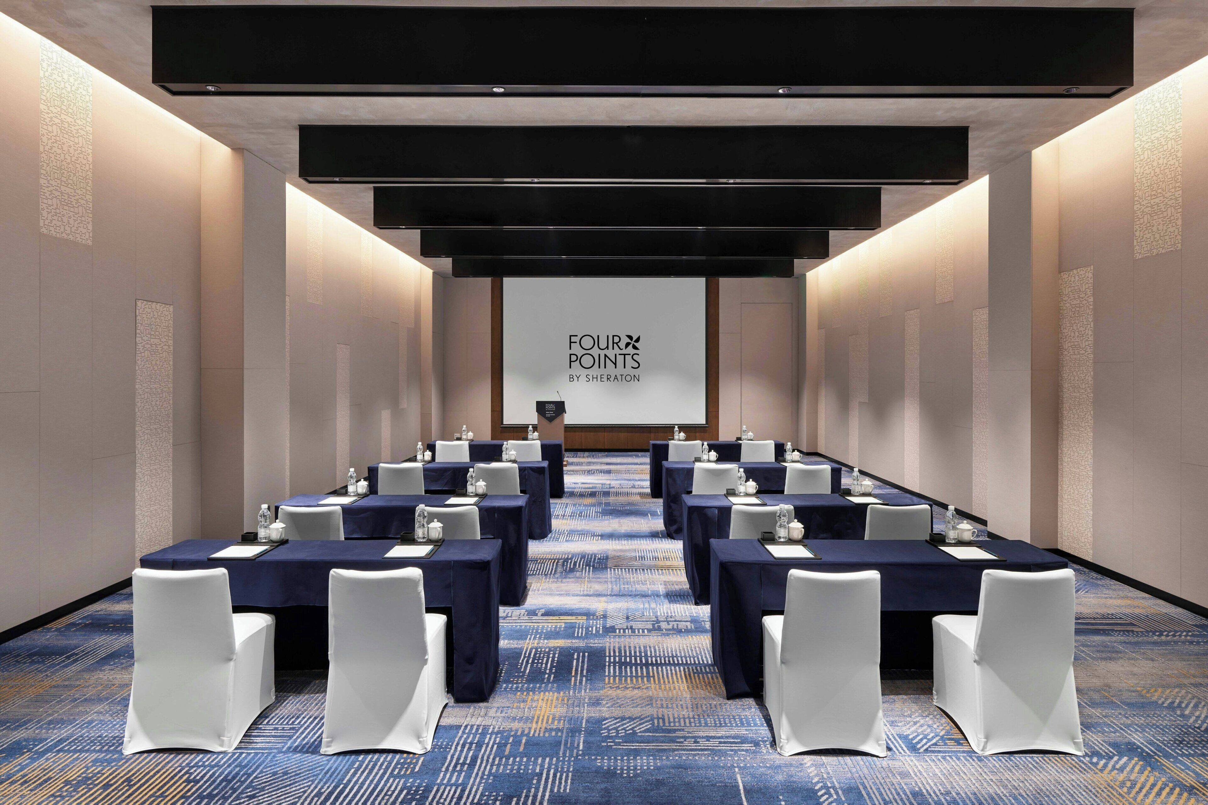 Ein Konferenzraum mit für eine Besprechung gedeckten Tischen, einem großen Bildschirm mit der Aufschrift "FOUR POINTS BY SHERATON" und einem Teppichboden.