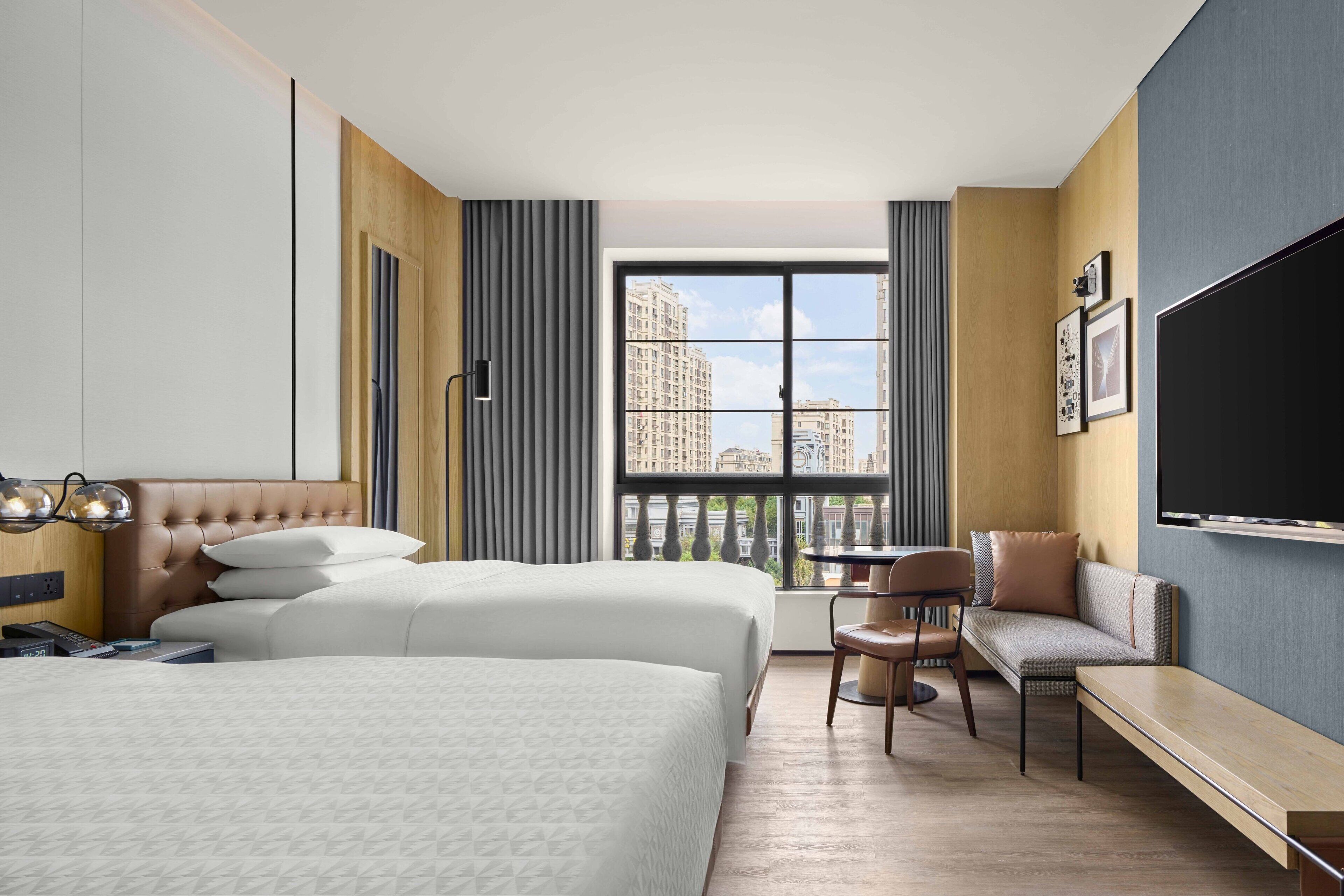 Ein Hotelzimmer mit zwei Betten, einem Schreibtisch, einem Stuhl, einem Fernseher und einem großen Fenster mit Blick auf die Stadt.