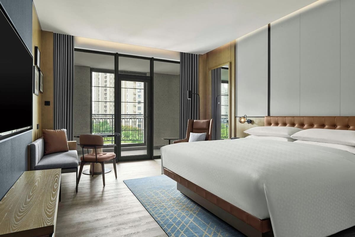 Une chambre d’hôtel avec un grand lit, un coin salon comprenant un canapé et des fauteuils, une petite table et une vue sur les bâtiments par la fenêtre.