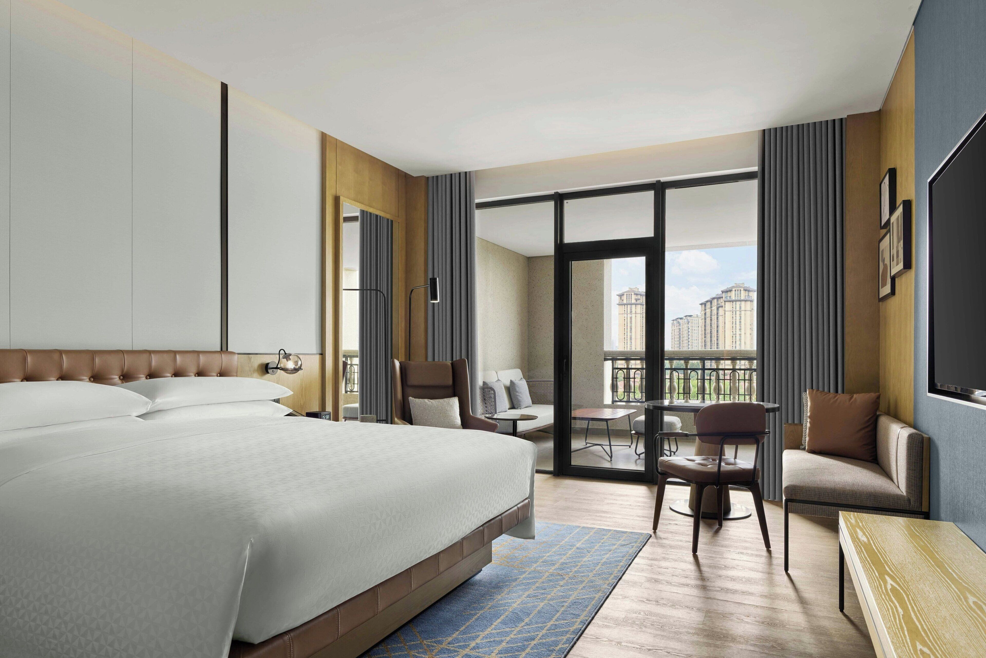A hotel room with a large bed, a seating area with a sofa and chairs, a balcony with a view of buildings, and a TV mounted on the wall.