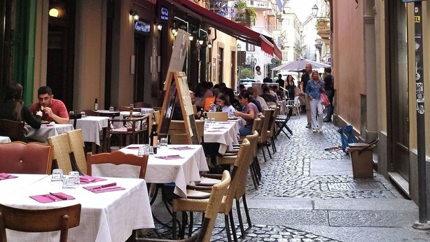 En brostensbelagt gade med caféer med udesiddepladser, et blåt banner og et skilt med 