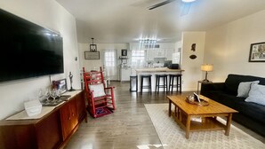 Un salón con televisión, una mesa de centro de madera, una silla roja, un sofá negro y una cocina al fondo.