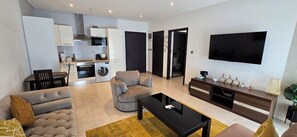 Een moderne woonkamer met een bank, fauteuil, salontafel en televisie. Achtergrond is een keuken met kasten, een fornuis en een wasmachine.