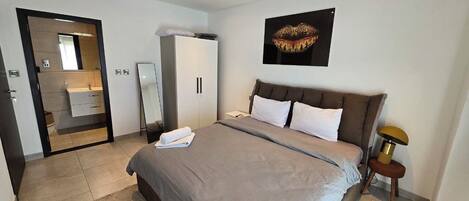 A hotel room with a bed, a wardrobe, a mirror, a bedside table with a lamp, and a painting on the wall.