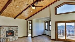 A spacious room with a stone fireplace, wooden flooring, and a large window offering a view of the mountains. - Above It all (Lake Arrowhead)
