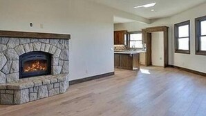 A spacious living area with a stone fireplace, wooden flooring, and a view of the kitchen in the background. - Above It all (Lake Arrowhead)