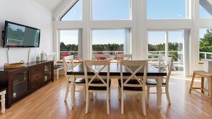 Un vaste espace dédié à la salle à manger, avec une grande table et des chaises, un téléviseur fixé au mur et de grandes fenêtres offrant une vue sur l’extérieur.