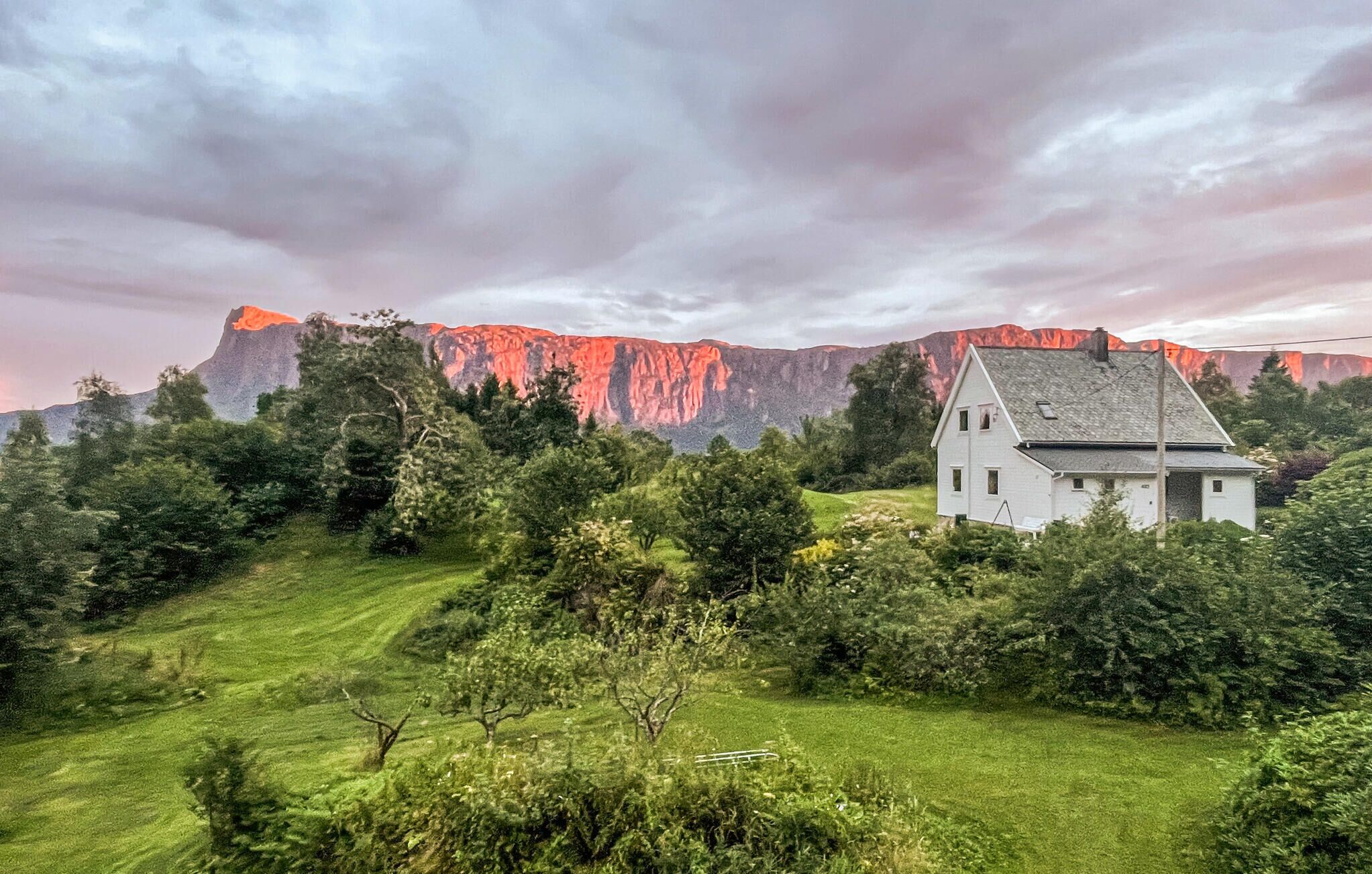 A white building with a sloped roof, surrounded by lush green grass and trees, with a mountain in the background.