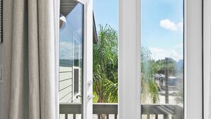 A view through a white door with a glass panel, leading to a balcony with a wooden railing, a palm tree, and a clear sky.