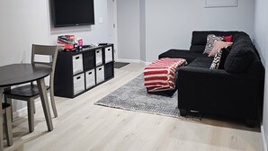 Una sala de estar moderna con una televisión de pantalla plana montada en la pared, un sofá negro con cojines estampados, una mesa de comedor con sillas y un mueble de almacenamiento con cajones.