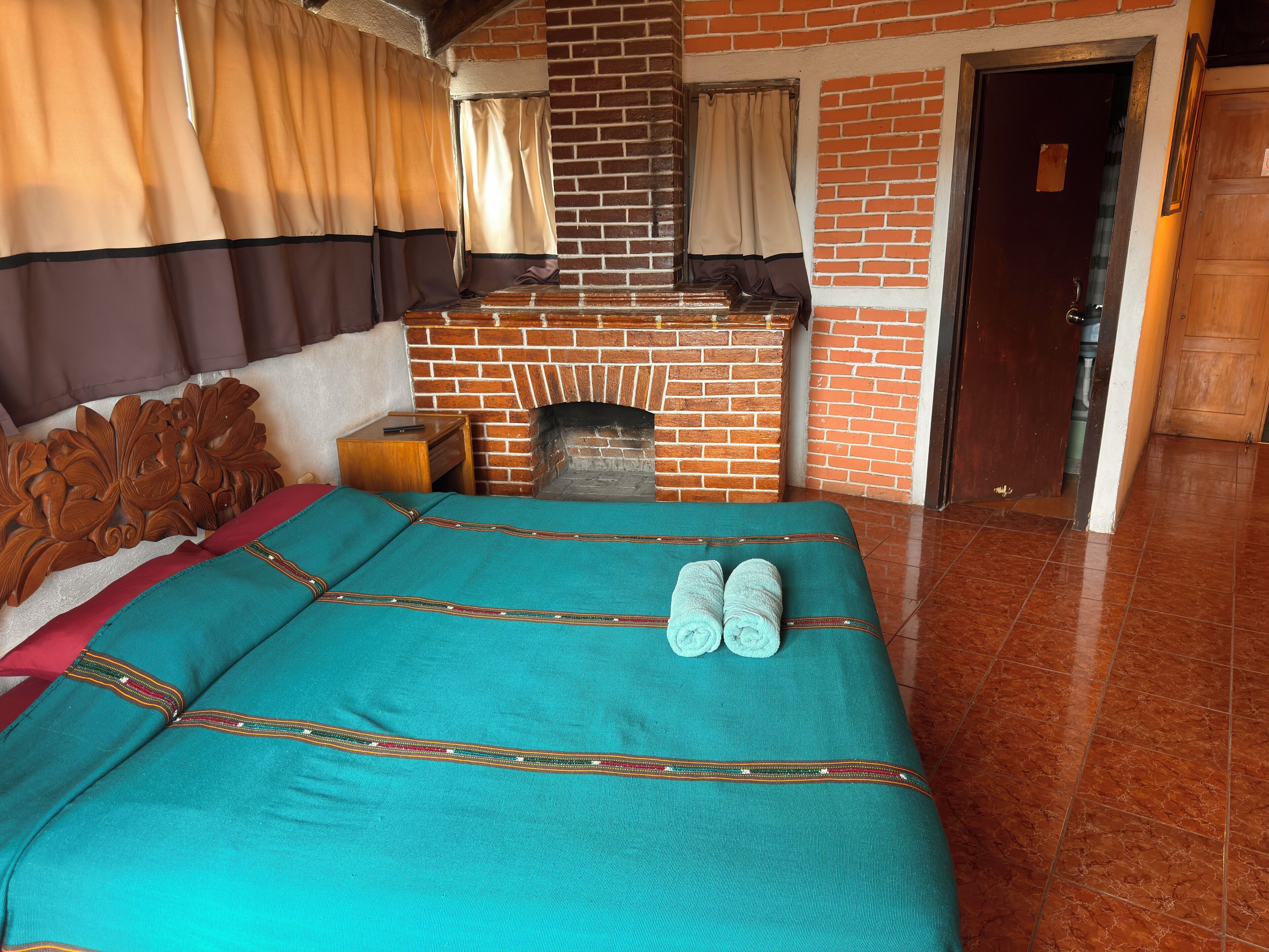 Una cama bien hecha con colcha color turquesa y un par de toallas, un mesita de noche de madera con un libro, y una chimenea de ladrillo al fondo.