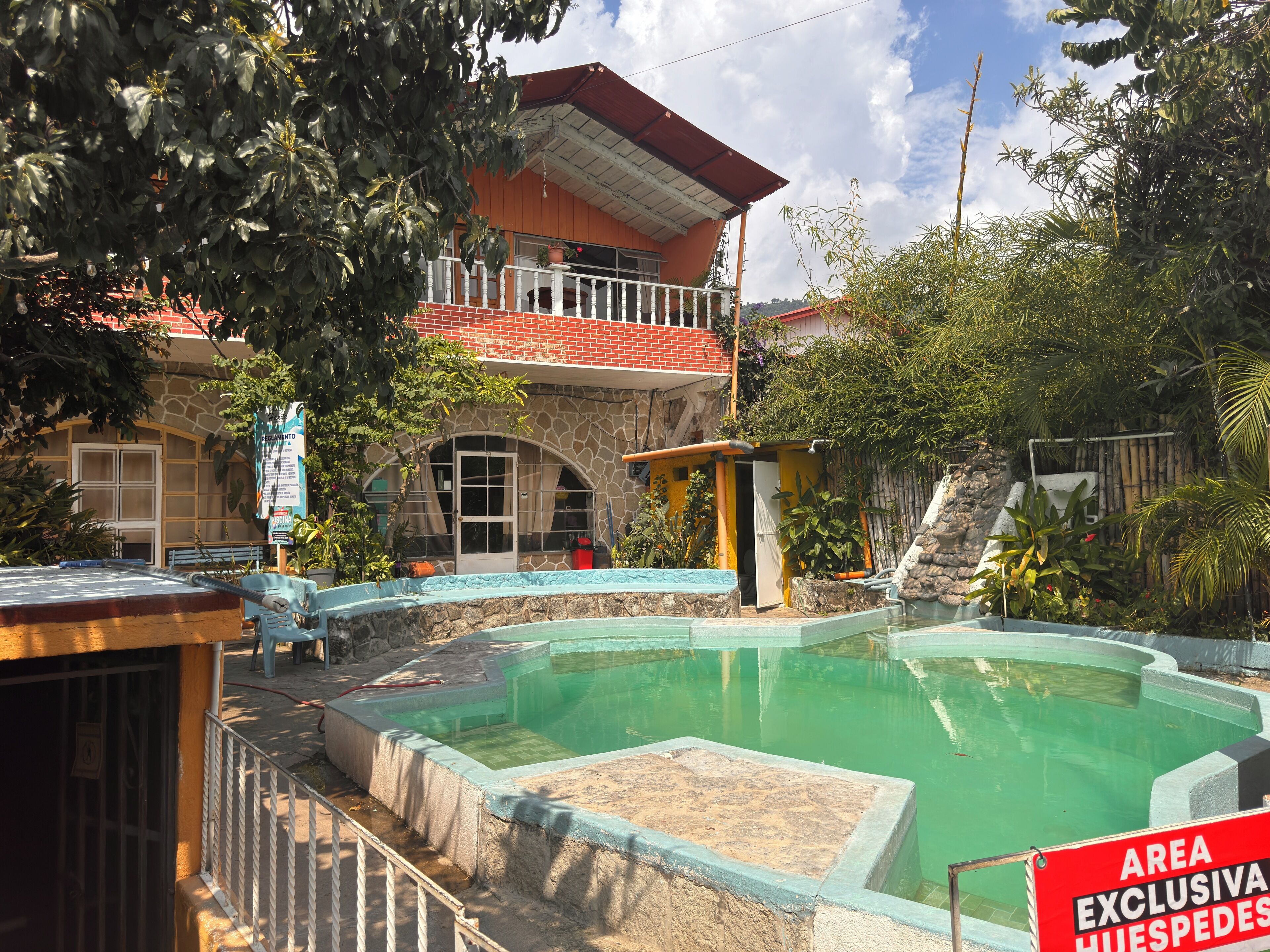 Un área de piscina con borde de piedra, una estructura pequeña y un edificio con techo rojo y balcón.