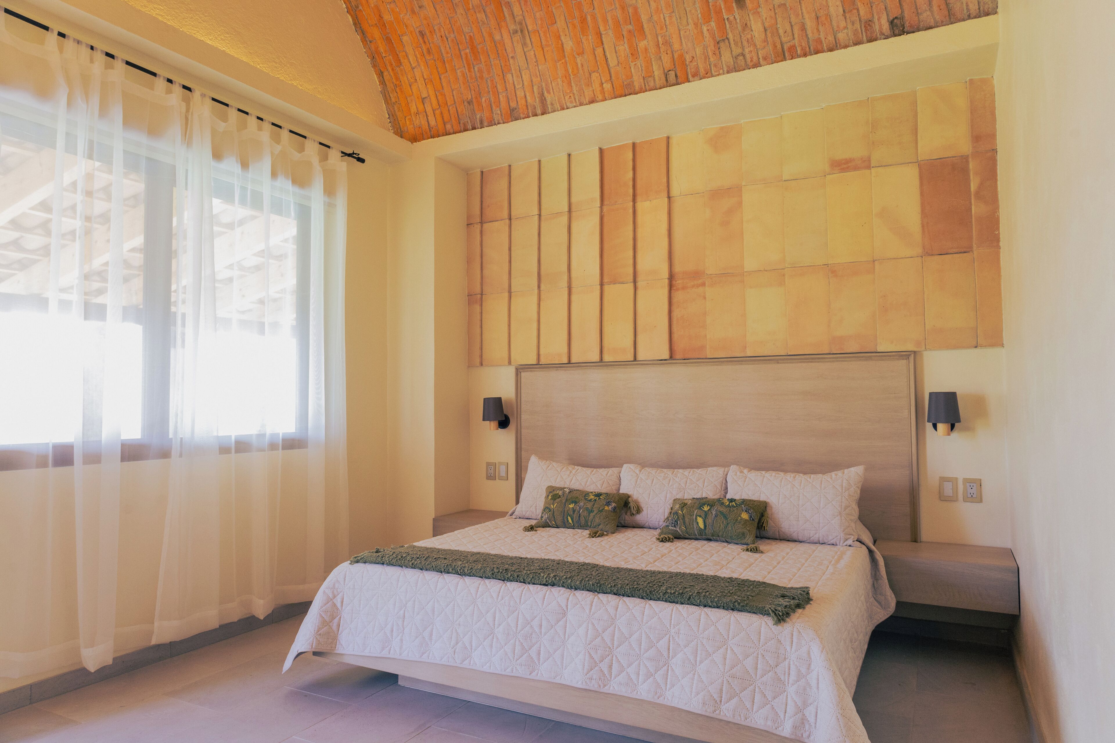 Un dormitorio con una cama grande, cabecera con estampado geométrico, dos lámparas de pared y cortinas de gasa.