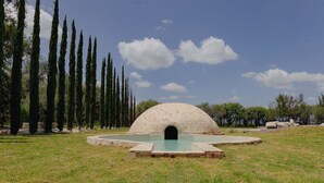 A large, dome-shaped structure with a central entrance, surrounded by a circular pool and a grassy area with tall trees. - Vid de Luna (San Miguel de Allende)