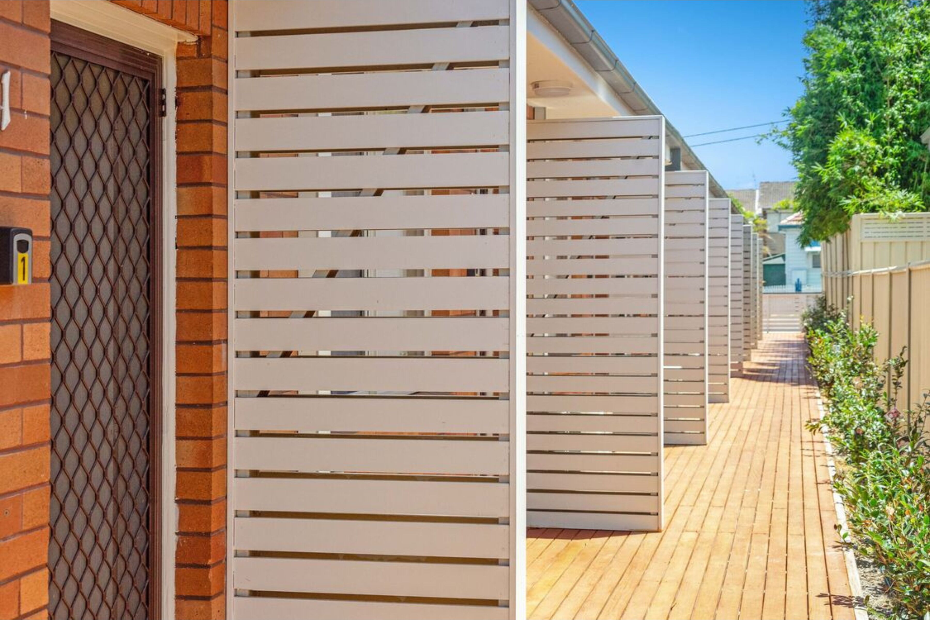 A row of white, horizontal slats along a wooden deck, with a brick wall and a metal gate on the left.