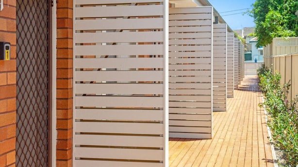 Una hilera de listones blancos horizontales a lo largo de una terraza de madera, con un muro de ladrillo y una verja metálica a la izquierda.