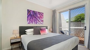 Habitación de hotel moderna con una cama grande, una mesita de noche con lámpara, un ventilador de techo y un ventanal con vistas al exterior.