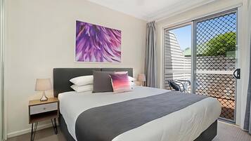A modern hotel room with a large bed, a nightstand with a lamp, a ceiling fan, and a large window with a view of the outside.