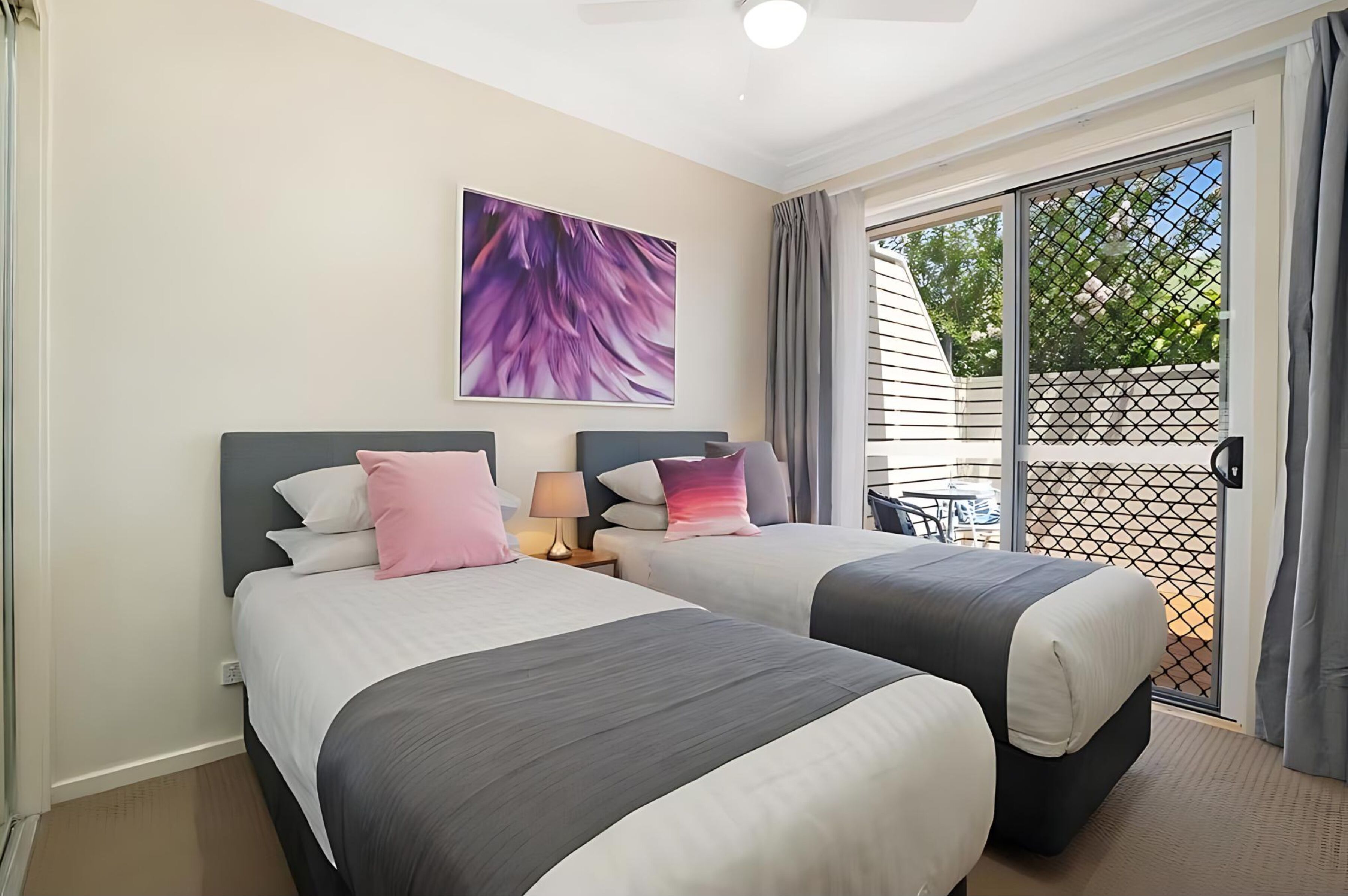 A hotel room with two beds, a ceiling fan, a large window with a view, and a table outside.