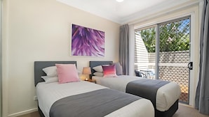 Habitación de hotel con dos camas, ventilador de techo, ventana grande con vistas y una mesa afuera.