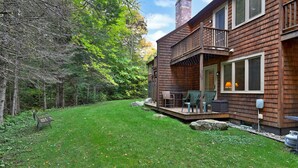 A wooden cabin with a deck, outdoor seating, and a grassy yard surrounded by trees.