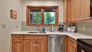 A kitchen with wooden cabinets, a stainless steel dishwasher, a window with a view of trees, and a coffee maker.